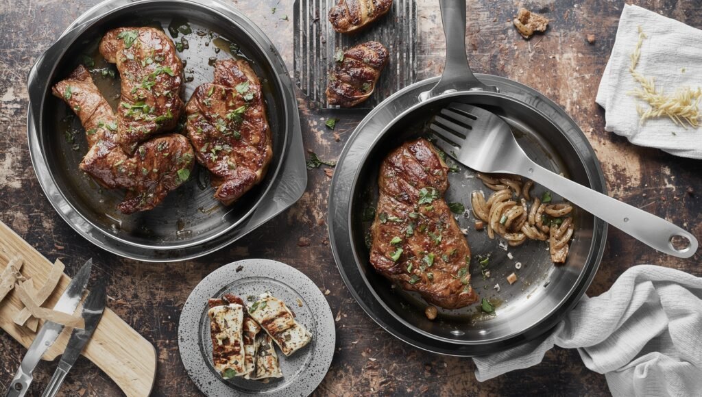 "All-Clad D5 stainless steel pans with seared steak, onions, and grilled cheese, showcasing premium non-toxic cookware tested by home cooks"